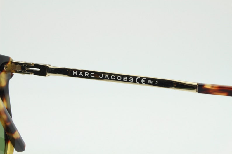 May include: Close-up of the arm of a pair of tortoiseshell sunglasses with gold metal arms. The arm is engraved with the text "MARC JACOBS CE EM 2".