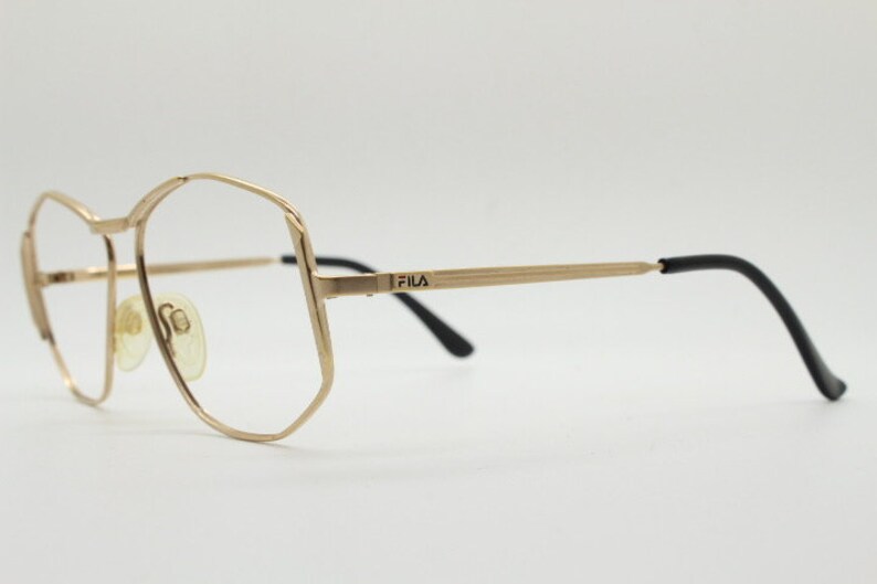 Puede incluir: Gafas de metal doradas con patillas de pl&aacute;stico negro. La montura tiene una forma geom&eacute;trica y el nombre de la marca "FILA" est&aacute; impreso en la patilla.