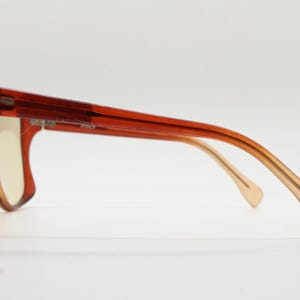 Op de afbeelding: Zijaanzicht van een bril met een dik, rechthoekig montuur in een doorschijnende roodbruine kleur. De pootjes lopen over van lichtbruin naar donkerder bruin. De glazen zijn lichtgeel. Het uiteinde van de pootjes heeft een klein, ge&euml;tst brillen-icoontje.