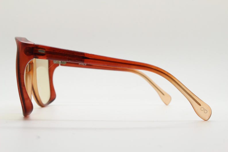 Op de afbeelding: Zijaanzicht van een bril met een dik, rechthoekig montuur in een doorschijnende roodbruine kleur. De pootjes lopen over van lichtbruin naar donkerder bruin. De glazen zijn lichtgeel. Het uiteinde van de pootjes heeft een klein, ge&euml;tst brillen-icoontje.