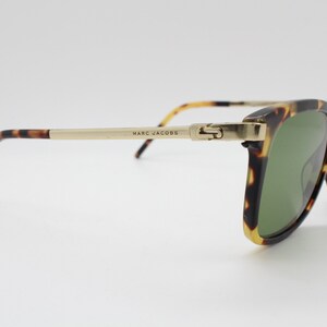 May include: Tortoiseshell sunglasses with gold metal accents and green lenses. The sunglasses have the brand name "MARC JACOBS" printed on the arm.