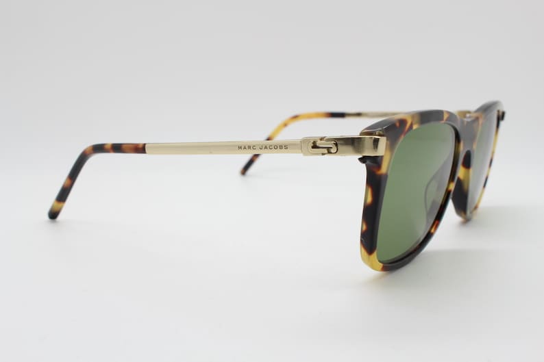 May include: Tortoiseshell sunglasses with gold metal accents and green lenses. The sunglasses have the brand name "MARC JACOBS" printed on the arm.