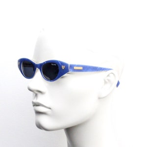 Op de afbeelding: Blauwe, ovale zonnebril met een hartvormig detail aan de zijkant. De zonnebril heeft een lichtblauw en wit marmerpatroon.