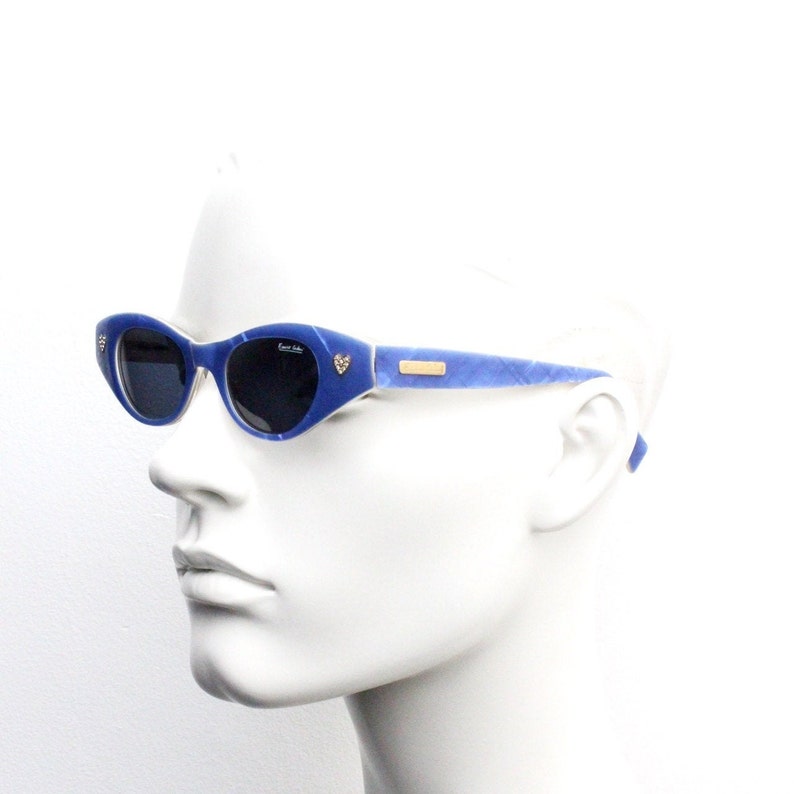 Op de afbeelding: Blauwe, ovale zonnebril met een hartvormig detail aan de zijkant. De zonnebril heeft een lichtblauw en wit marmerpatroon.