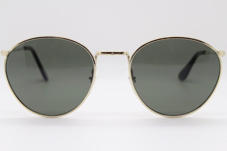 Pu&ograve; includere: Occhiali da sole rotondi con montatura in metallo dorato e lenti verde scuro. Il design classico presenta un doppio ponte e aste sottili. Questi occhiali offrono un look elegante per l'uso quotidiano.
