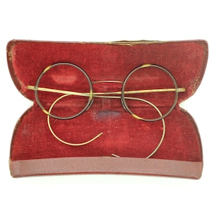 Peut inclure: Lunettes rondes vintage avec des montures à motif écaille de tortue, posées dans un étui ouvert doublé de velours rouge. Les lunettes ont de fines branches et un pont en métal doré. L'étui est de couleur rouge foncé.