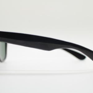 Puede incluir: Gafas de sol negras con un dise&ntilde;o cl&aacute;sico. Las monturas son brillantes y las lentes son de un tono oscuro. Las gafas de sol est&aacute;n sobre un fondo blanco, mostrando su perfil elegante.