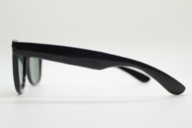 Puede incluir: Gafas de sol negras con un dise&ntilde;o cl&aacute;sico. Las monturas son brillantes y las lentes son de un tono oscuro. Las gafas de sol est&aacute;n sobre un fondo blanco, mostrando su perfil elegante.