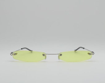 Könnte beinhalten: Randlose Sonnenbrille mit hellgelber Tönung. Die ovalen Gläser werden von einem dünnen silbernen Metallrahmen zusammengehalten. Die Bügel sind dunkelgrau.