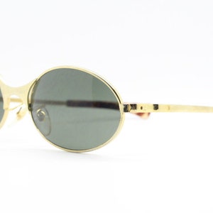 Puede incluir: Gafas de sol ovaladas doradas con lentes verde oscuro. Las gafas tienen un fino marco de metal y un puente curvado.