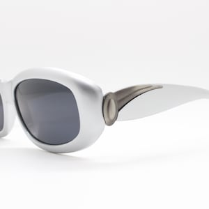 Può includere: Occhiali da sole argentati con lenti scure. Gli occhiali hanno una montatura arrotondata e oversize e una finitura metallica. Le aste sono curve e presentano un accento metallico decorativo vicino alle cerniere. Su sfondo bianco.