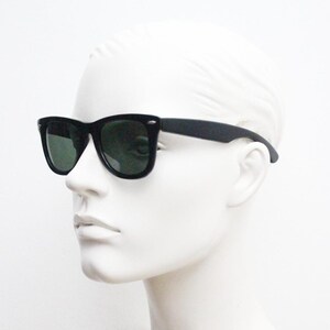 Puede incluir: Gafas de sol negras con lentes verde oscuro. Las gafas tienen una montura rectangular cl&aacute;sica y se muestran en una cabeza de maniqu&iacute; blanca. Las patillas son rectas.