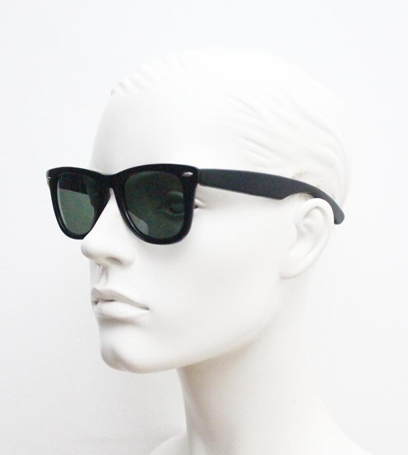 Puede incluir: Gafas de sol negras con lentes verde oscuro. Las gafas tienen una montura rectangular cl&aacute;sica y se muestran en una cabeza de maniqu&iacute; blanca. Las patillas son rectas.