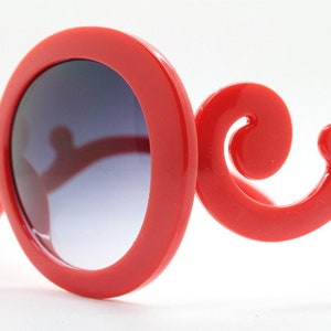 May include: Red plastic sunglasses with round lenses and a unique, ornate, swirl design on the arms.