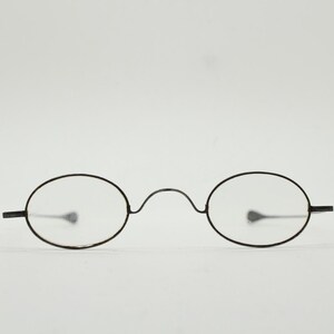 Puede incluir: Gafas de forma ovalada con una fina montura de metal negro. Las lentes son transparentes y el puente de las gafas tiene una curva sutil. Las patillas se extienden horizontalmente desde las lentes. Un diseño de gafas clásico.