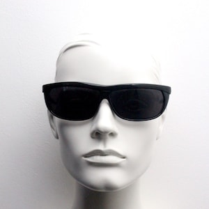 Könnte beinhalten: Schwarze Sonnenbrille mit dunklen Gläsern. Die Sonnenbrille hat eine rechteckige Form mit einem leicht gebogenen oberen Rahmen. Das Bild ist eine Nahaufnahme der Sonnenbrille auf einem Schaufensterkopf vor weißem Hintergrund.