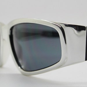 Könnte beinhalten: Silberne und schwarze Sonnenbrille mit einem schlanken, modernen Design. Die Sonnenbrille hat einen gebogenen Rahmen und dunkle Gläser.