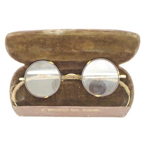 Peut inclure: Lunettes rondes vintage avec une monture dorée et des verres clairs, présentées dans un étui ouvert doublé de velours marron. L'étui est rectangulaire avec un couvercle à charnière. Le texte "36 Manchester Road, Bradford" est visible.