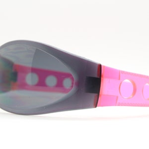 May include: Gray and pink sunglasses with a sporty design. The lenses are tinted gray and the frame has a pink accent with holes.