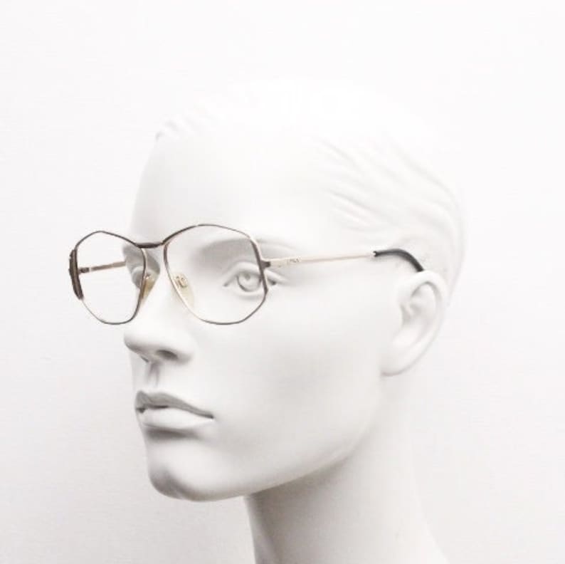 Puede incluir: Un maniqu&iacute; con gafas de montura dorada con lentes octogonales. Las gafas tienen un puente de nariz negro y patillas negras.
