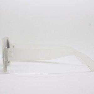 May include: A pair of white plastic sunglasses with a rounded frame and a dark lens. The sunglasses are shown in profile view.