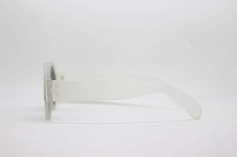 May include: A pair of white plastic sunglasses with a rounded frame and a dark lens. The sunglasses are shown in profile view.