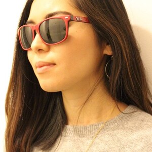 May include: A woman with long brown hair wearing red sunglasses with the Ray-Ban logo on the side. She is wearing a gray sweater and a gold necklace.