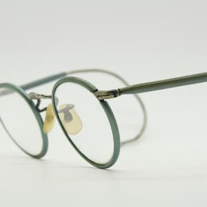 May include: Round eyeglasses featuring sage green circular rims and matching thin temples. The frame includes a metallic bridge, clear lenses, and pale yellow nose pads. The design incorporates curved ear pieces for a secure fit around the ears.