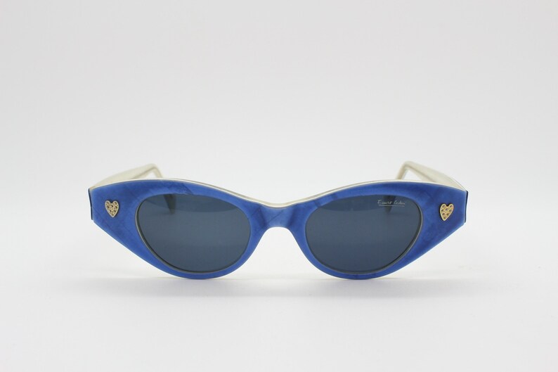 Op de afbeelding: Blauwe cat-eye zonnebril met een wit montuur en gouden hart accenten. De zonnebril heeft een "Fendi Italia" label op de rechter lens.