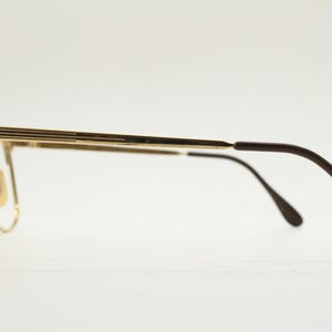 Puede incluir: Gafas de metal dorado con patillas de pl&aacute;stico marr&oacute;n. Las gafas tienen una montura rectangular y un puente de metal fino.