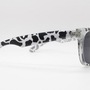 May include: Clear, cow print sunglasses with black lenses. The arms of the sunglasses are white with a black cow print pattern.