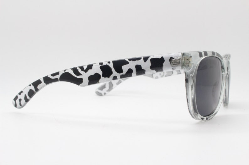 May include: Clear, cow print sunglasses with black lenses. The arms of the sunglasses are white with a black cow print pattern.