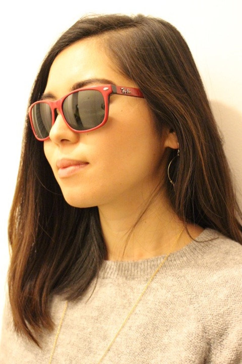 May include: A woman with long brown hair wearing red sunglasses with the Ray-Ban logo on the side. She is wearing a gray sweater and a gold necklace.