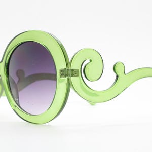 May include: Translucent lime green sunglasses featuring large circular frames and ornate, swirling scrollwork details on the temples. The lenses are dark and gradient, transitioning from a deeper tint at the top to a lighter shade at the bottom.