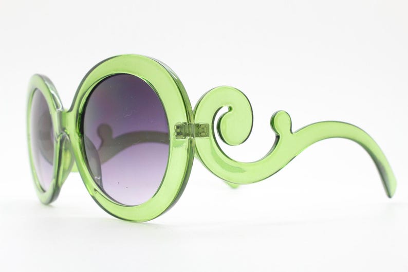 May include: Translucent lime green sunglasses featuring large circular frames and ornate, swirling scrollwork details on the temples. The lenses are dark and gradient, transitioning from a deeper tint at the top to a lighter shade at the bottom.
