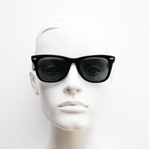 Puede incluir: Gafas de sol negras con lentes verde oscuro. Las gafas de sol tienen un dise&ntilde;o de montura rectangular cl&aacute;sico. La imagen muestra las gafas de sol en una cabeza de maniqu&iacute; blanca, destacando la forma y el estilo de las gafas.