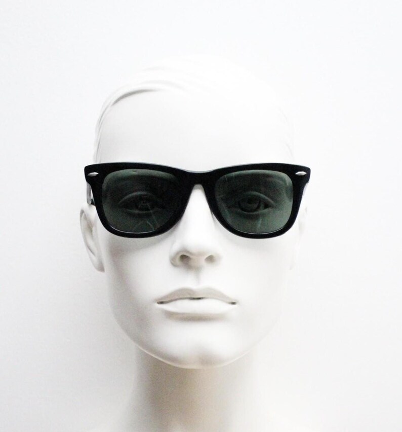 Puede incluir: Gafas de sol negras con lentes verde oscuro. Las gafas de sol tienen un dise&ntilde;o de montura rectangular cl&aacute;sico. La imagen muestra las gafas de sol en una cabeza de maniqu&iacute; blanca, destacando la forma y el estilo de las gafas.