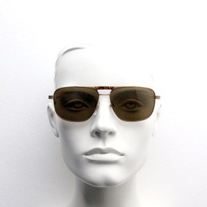 May include: A gold-framed pair of aviator sunglasses with brown lenses. The sunglasses are on a white mannequin head.