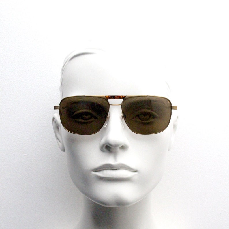 May include: A gold-framed pair of aviator sunglasses with brown lenses. The sunglasses are on a white mannequin head.
