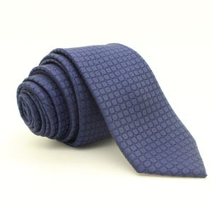 May include: Rolled-up blue tie with a repeating geometric pattern. The tie is made of a textured fabric and is displayed against a white background. The tie is a classic accessory.