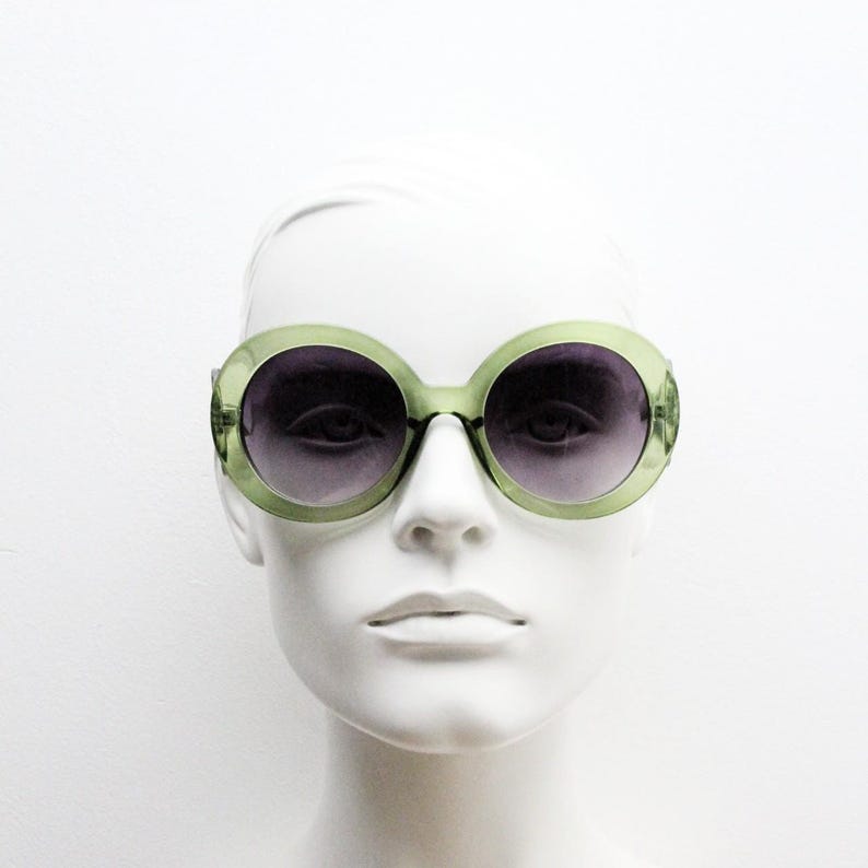 May include: Oversized round sunglasses featuring translucent light green plastic frames and dark gradient lenses. The circular frames have a thick rim design that surrounds the dark tinted lenses, providing a bold look on a white mannequin head.