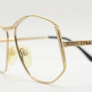 Puede incluir: Gafas de metal doradas con montura de forma geom&eacute;trica y la palabra "FILA" impresa en el lateral.