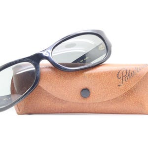 May include: Black sunglasses with a rounded frame rest on a brown leather case. The case has a button closure and the word "Polaroid" is embossed on the front. The sunglasses have a dark tint and are marked "Made in USA".