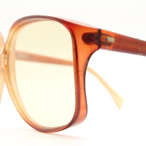 Op de afbeelding: Vintage oversized bril met een warme amberkleur. De monturen zijn een doorschijnend oranje-bruin en de lenzen hebben een gele tint. De pootjes hebben een vergelijkbare kleur, met een lichte curve. Een klassiek accessoire voor een retro look.