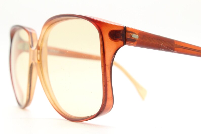 Op de afbeelding: Vintage oversized bril met een warme amberkleur. De monturen zijn een doorschijnend oranje-bruin en de lenzen hebben een gele tint. De pootjes hebben een vergelijkbare kleur, met een lichte curve. Een klassiek accessoire voor een retro look.