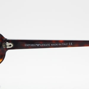 Op de afbeelding: Emporio Armani zonnebril met een montuur van schildpad en donkere glazen. De tekst "EMPORIO ARMANI MADE IN ITALY CE" staat op de poot. De zonnebril heeft een klassieke ovale vorm.