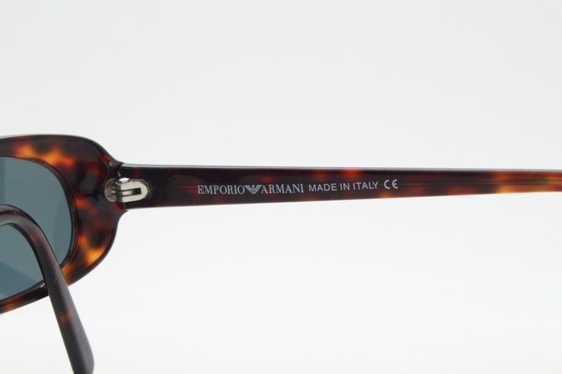 Op de afbeelding: Emporio Armani zonnebril met een montuur van schildpad en donkere glazen. De tekst "EMPORIO ARMANI MADE IN ITALY CE" staat op de poot. De zonnebril heeft een klassieke ovale vorm.