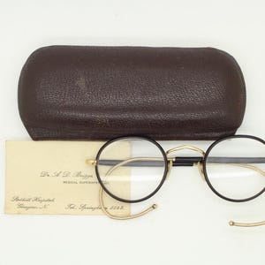 May include: Vintage round eyeglasses with black frames and gold-toned accents, resting on a white surface. Includes a brown leather case and a cream-colored card with text. The glasses have curved temples.