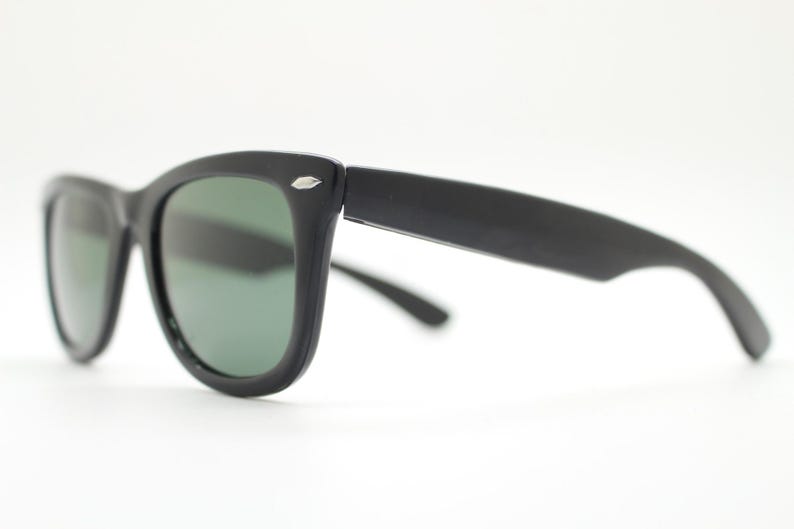 Puede incluir: Gafas de sol negras con un dise&ntilde;o cl&aacute;sico y retro. Las monturas son gruesas y mate, con un peque&ntilde;o acento plateado en las patillas. Las lentes tienen un tinte verde, ofreciendo un contraste elegante. Un accesorio atemporal.
