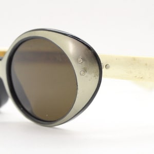 Może przedstawiać: Okulary przeciwsłoneczne vintage z srebrną i czarną oprawką i brązowymi szkłami. Okulary mają kształt kociego oka i są w dobrym stanie.